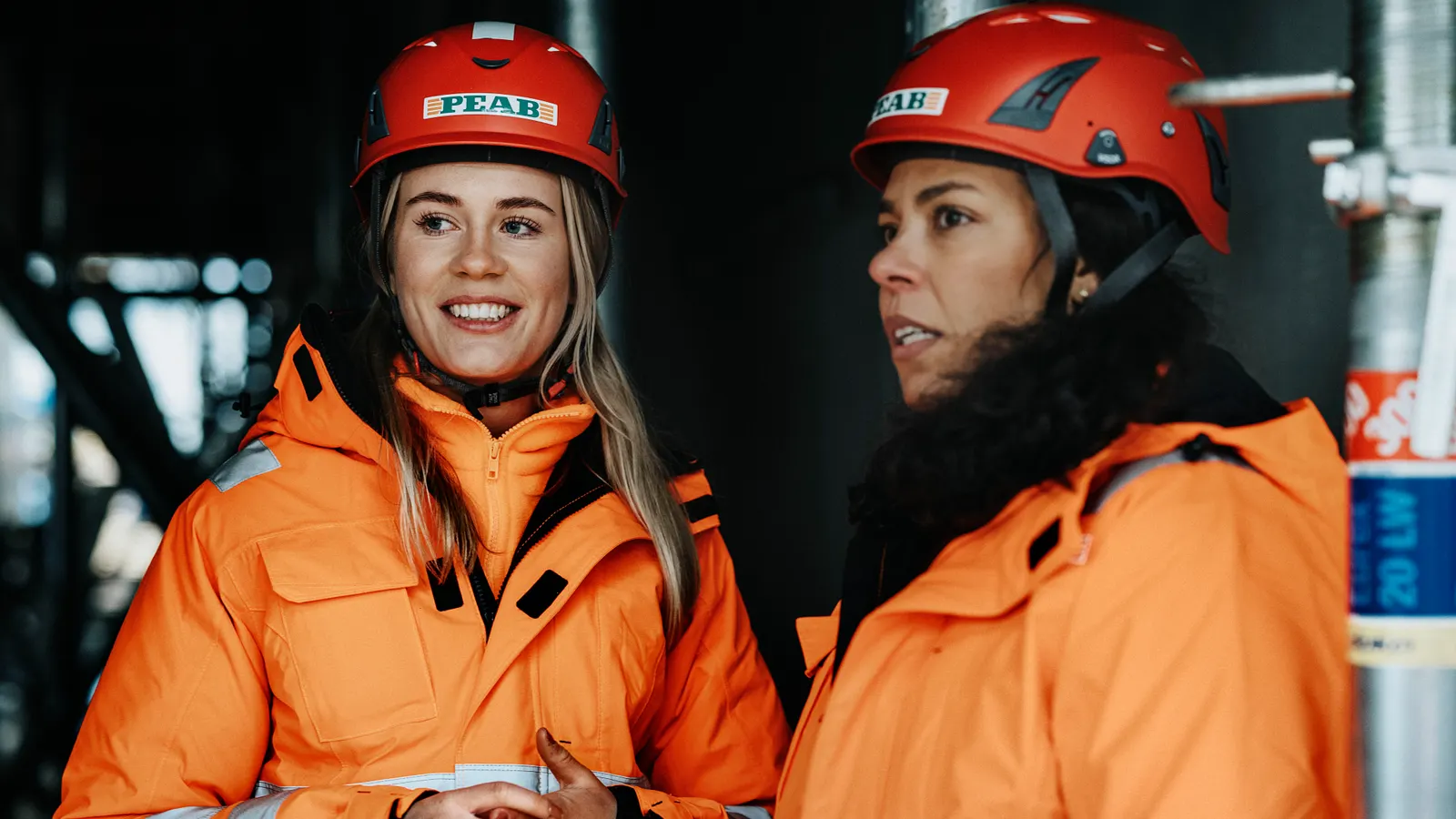 Two Peab construction workers in orange high-visibility jackets and red Peab helmets on site